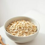 Can You Eat Oats Safely With Coeliac Disease? Bowl of certified gluten free oats with a wooden spoon and oat stalks